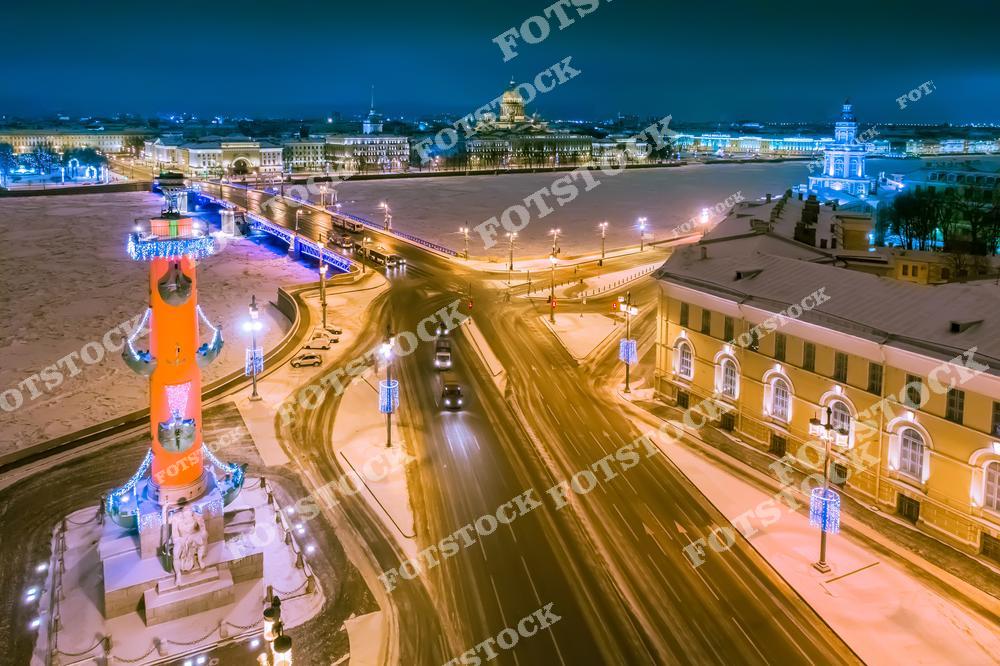 На фотографии запечатлена Стрелка Васильевского острова, украшенная вечерней подсветкой. Ростральная колонна, сверкающая огнями, выделяется на фоне замерзшей Невы. На заднем плане видны Дворцовый мост и здание Адмиралтейства. Заснеженные улицы и свет фонарей создают атмосферу зимнего праздника в Санкт-Петербурге.